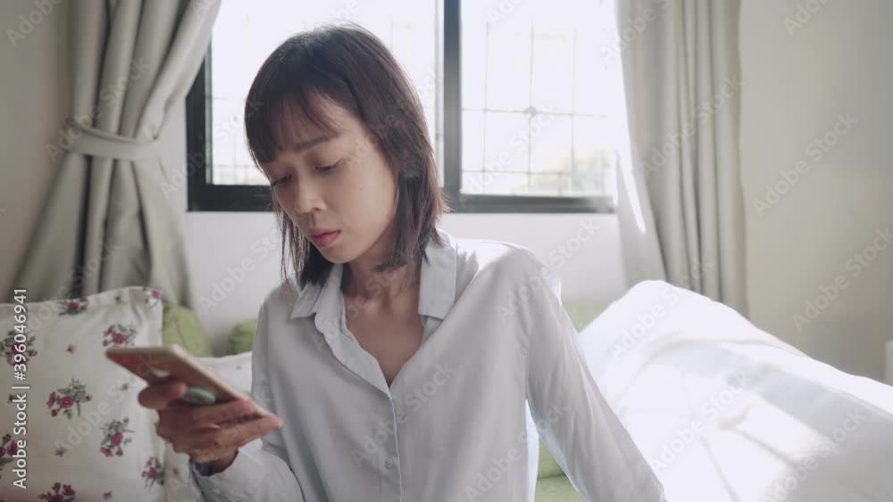 Asian woman using smartphone sitting down on couch at home living room, on bright sunny day, sending text message chatting, online communications technology, single female using dating application Asian woman using smartphone sitting down on couch at home living room, on bright sunny day, sending text message chatting, online communications technology, single female using dating application