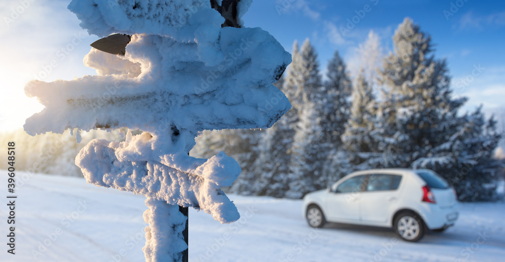 Fototapeta premium Snow covered signboard with wooden arrows on a background of a car on a road. Winter travel 