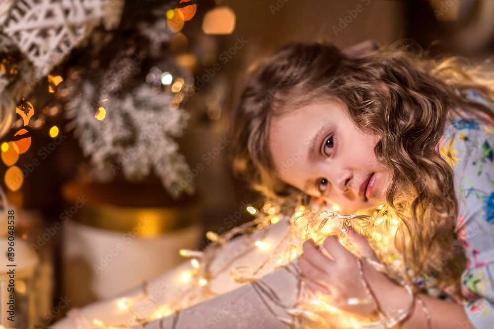 A beautiful girl with curly long hair is sitting near a Christmas tree. Holding a garland of Christmas lights. A beautiful girl with curly long hair is sitting near a Christmas tree. Holding a garland of Christmas lights.