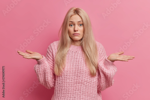 Uncertain blonde woman spreads palms and stands doubtful indoor cannot make choice between two options wears knitted warm sweater isolated over pink background. Doubtful female model shrugs shoulders