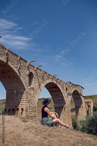 woman traveler Explorer in a black hat sit next to the destroyed old stone bridge in the summer photo