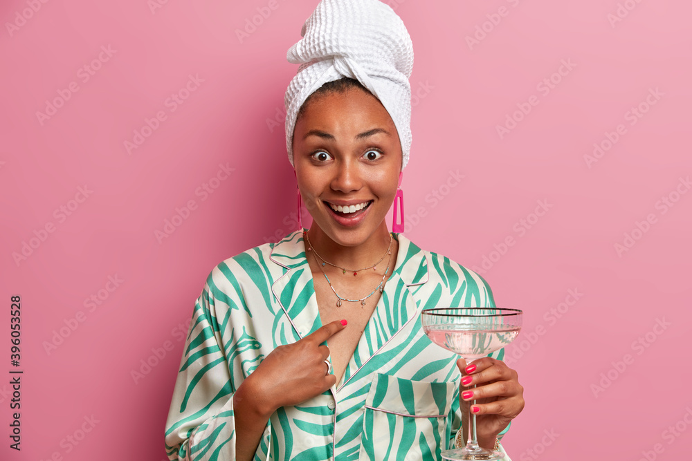Surprised cheerful young woman points at herself smiles broadly has white perfect teeth holds martini glass wears wrapped towel on washed head and dressing gown spends free time on pajama party. Surprised cheerful young woman points at herself smiles broadly has white perfect teeth holds martini glass wears wrapped towel on washed head and dressing gown spends free time on pajama party.