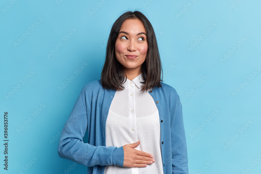 Pleased cheerful Asian woman keeps hand on belly feels full after delicious dinner dressed casually stands thoughtful against blue background. Happy young female finds out about her pregnancy Pleased cheerful Asian woman keeps hand on belly feels full after delicious dinner dressed casually stands thoughtful against blue background. Happy young female finds out about her pregnancy