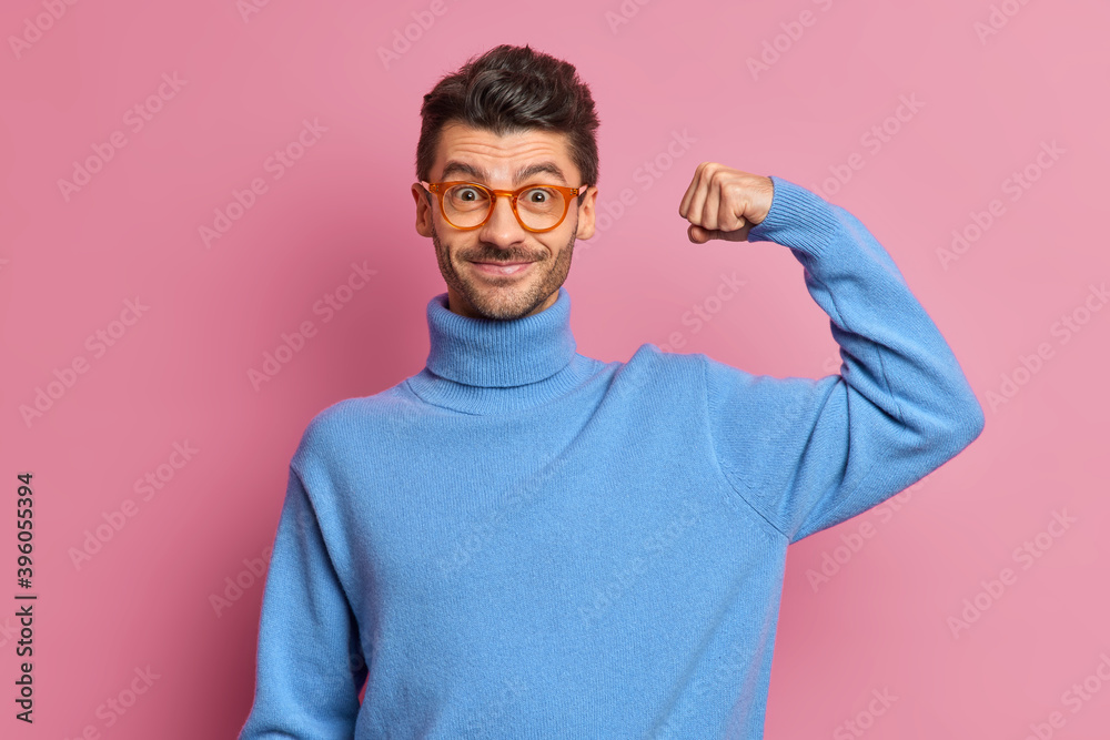 Self confident cheerful unshaven man raises arm and shows muscles feels power wears optical glasses and blue turtleneck demonstrates his power isolated over pink wall. Results after training in gym Self confident cheerful unshaven man raises arm and shows muscles feels power wears optical glasses and blue turtleneck demonstrates his power isolated over pink wall. Results after training in gym