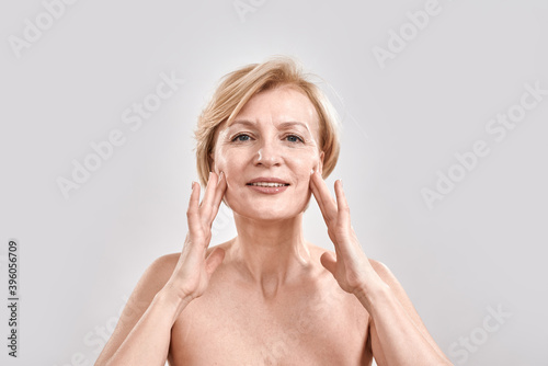 Portrait of beautiful middle aged woman satisfied with skincare treatment. She is touching her face  looking at camera while posing isolated against grey background. Beauty concept
