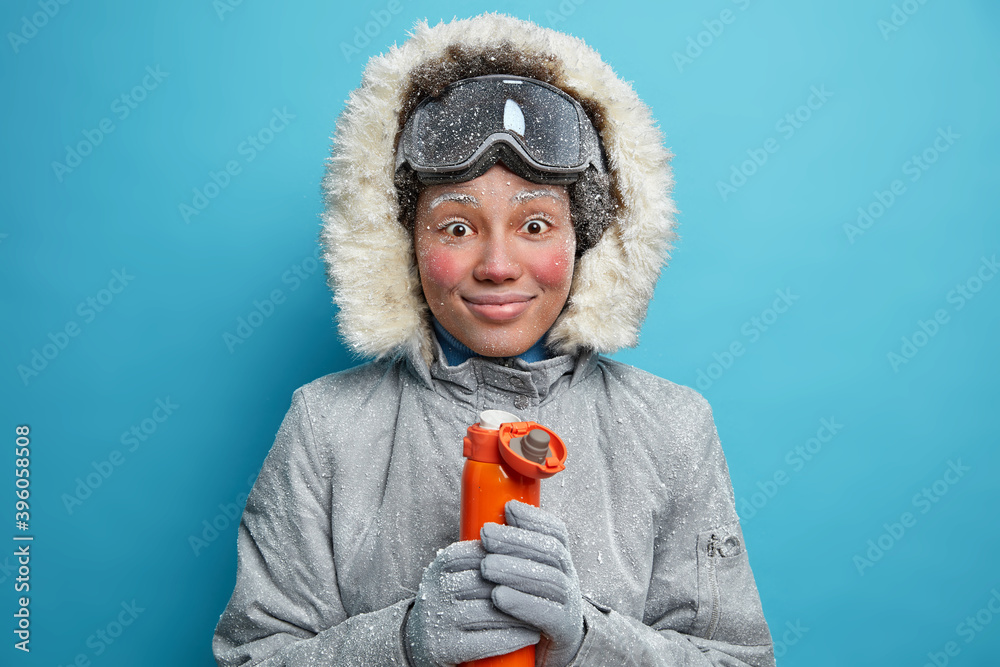 Cheerful frozen woman with red face during frost smiles pleasantly has coffee break after snowboarding activity wears warm winter clothes holds thermos with hot beverage poses against blue background Cheerful frozen woman with red face during frost smiles pleasantly has coffee break after snowboarding activity wears warm winter clothes holds thermos with hot beverage poses against blue background
