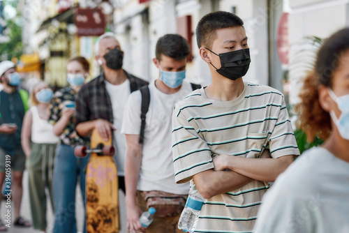Young asian guy wearing mask waiting  standing in line with other people  respecting social distancing to collect his takeaway order from the pickup point during coronavirus lockdown