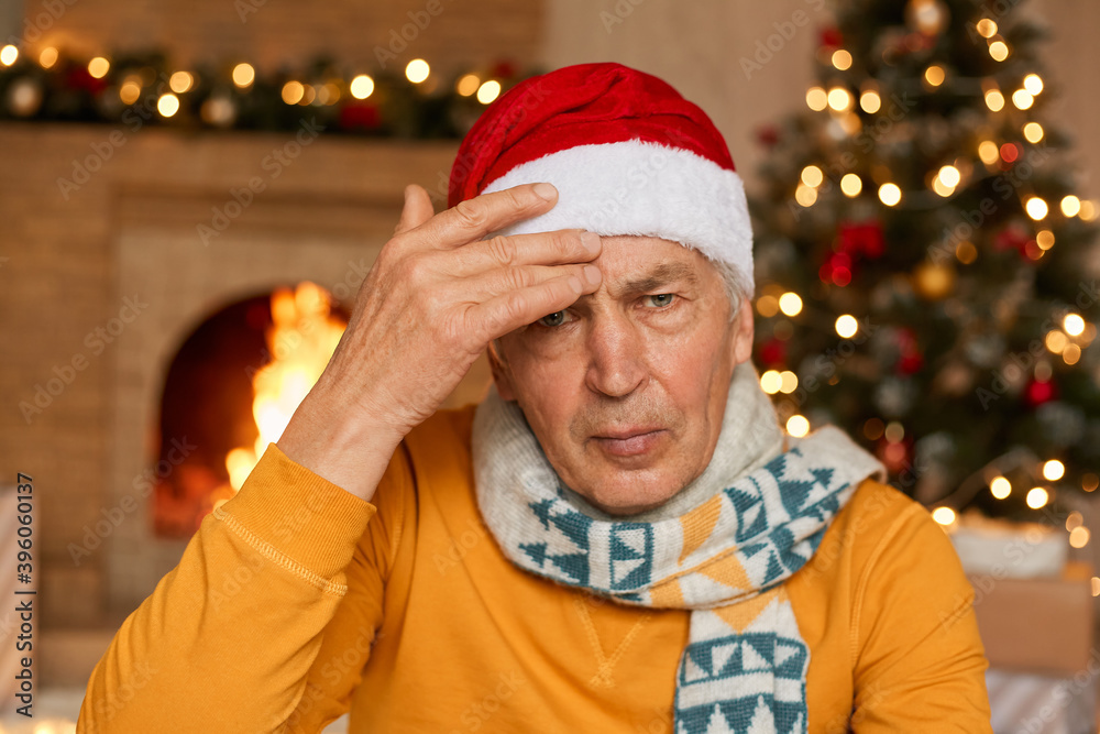 Tired unhappy annoyed old man being sick on Christmas eve, suffering from headache, keeping hand on forehead, wearing yellow shirt and santa hat, looks at camera with upset expression. Tired unhappy annoyed old man being sick on Christmas eve, suffering from headache, keeping hand on forehead, wearing yellow shirt and santa hat, looks at camera with upset expression.