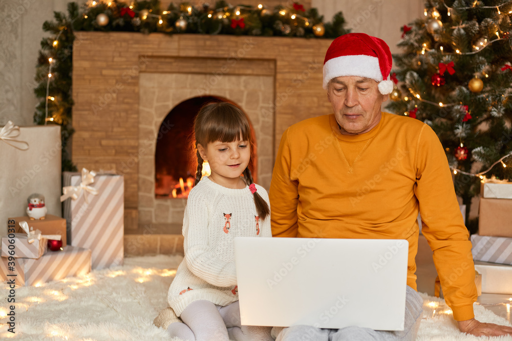 Charming little girl with grandfather sitting and using digital laptop during Christmas Eve, sitting on floor on soft carper near fir tree and fireplace, family looking concentrated at screen. Charming little girl with grandfather sitting and using digital laptop during Christmas Eve, sitting on floor on soft carper near fir tree and fireplace, family looking concentrated at screen.
