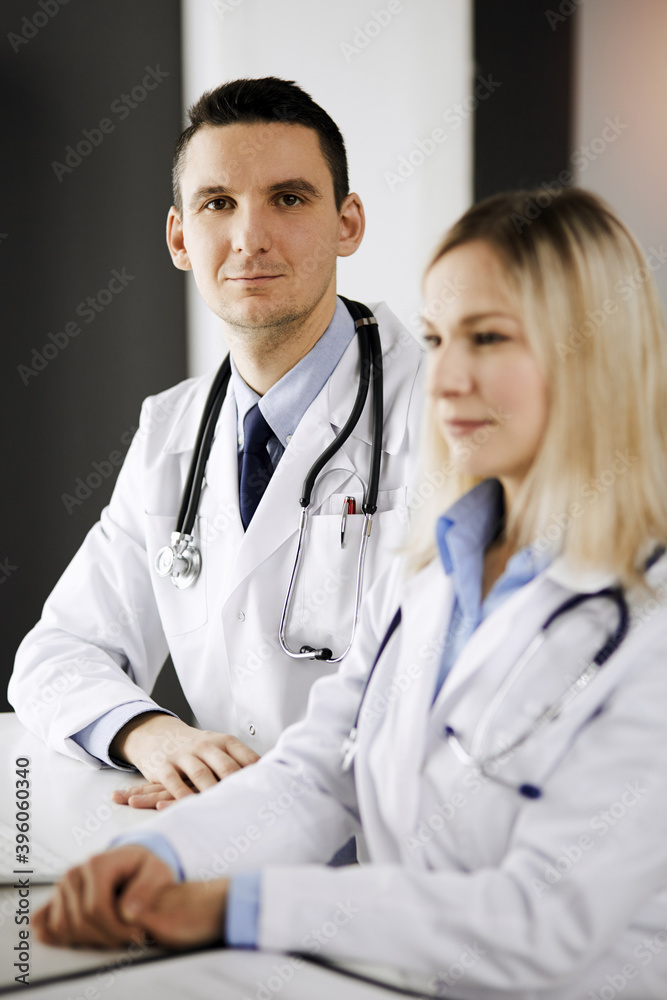 Two doctors colleagues discussing current disease therapy while sitting at working place in sunny clinic. Team work in hospital Two doctors colleagues discussing current disease therapy while sitting at working place in sunny clinic. Team work in hospital