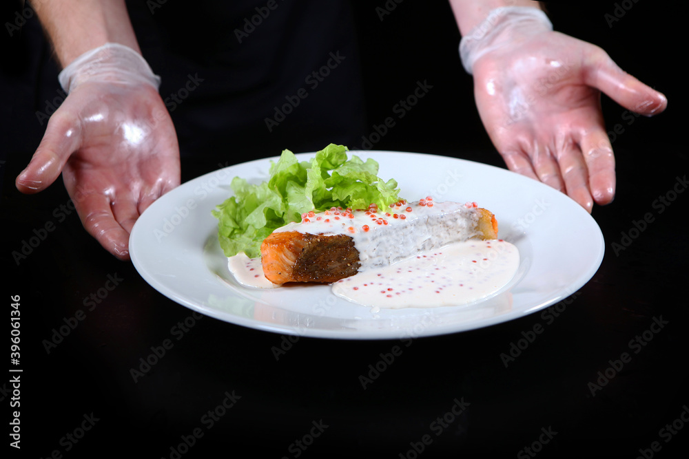 The cook serves a prepared fish dish. Salmon in creamy caviar sauce on a white plate. Lettuce leaves. Photo on a black background. Unrecognizable person.
