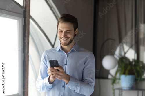 Positive smiling young man office worker standing by window at workplace reading good news from cellphone screen, having pleasant chat conversation in messenger, watching funny video send by colleague