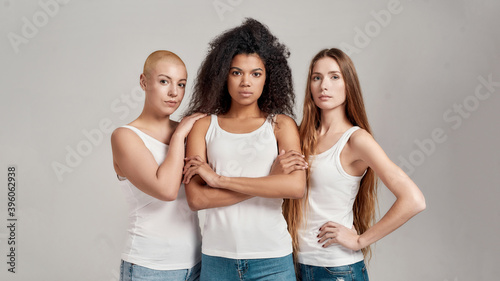 Three confident young diverse women wearing white shirts looking at camera while posing together isolated over grey background
