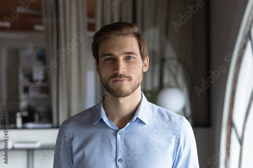 Headshot portrait of calm confident handsome millennial businessman qualified professional specialist standing in modern office looking at camera able to help consult client by video link using webcam photo