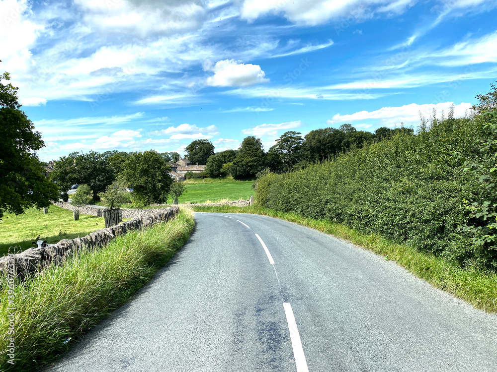 View along, Gledstone Road, with high hedgerow, dry stone walls, and buildings in the distance in, Martons Both, Skipton, UK