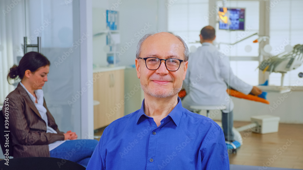 Fototapeta premium Senior man looking at camera while doctor examining patient in background. Elderly male smiling on webcam sitting on chair in waiting room of stomatological clinic, assistant typing on pc