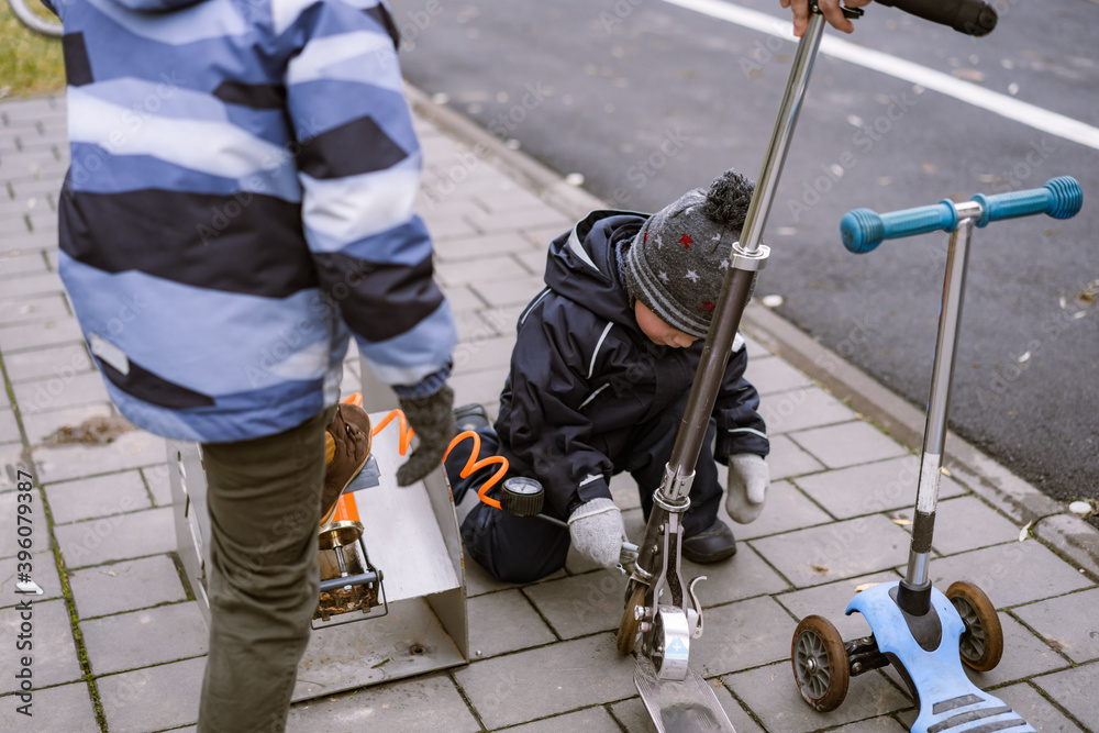 little boys pumping up a tire of their kick scooters with foot pump self service in the street, sitting on pavement