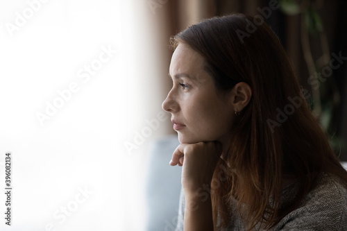 Pensive young woman looking in distance at window, thinking or life problems, suffering from loneliness or depressive mood, regretting mistakes, having psychological mental problems, copy space.
