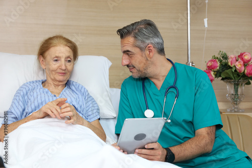 Doctor or assistance staff talking with an elder patient woman while using smart tablet and sitting on bed in the hospital