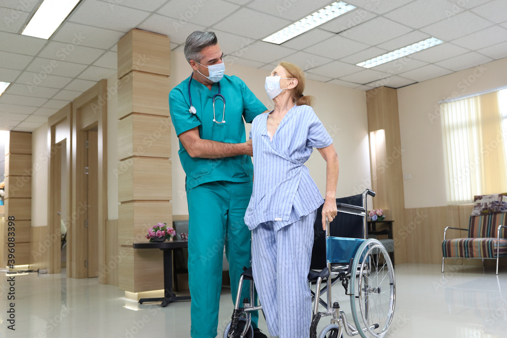 Fototapeta premium Doctor or assistance staff wear face mask and help or assist an elder patient woman while try to standing or walking or sitting on wheelchair in the hospital, eldercare concept