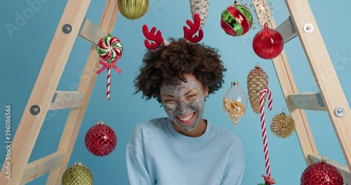 People winter holidays and beauty treatments concept. Joyful ethnic woman applies clay mask on face preapares for New Year corporative party laughs happily wears red reindeer antlers poses at home photo