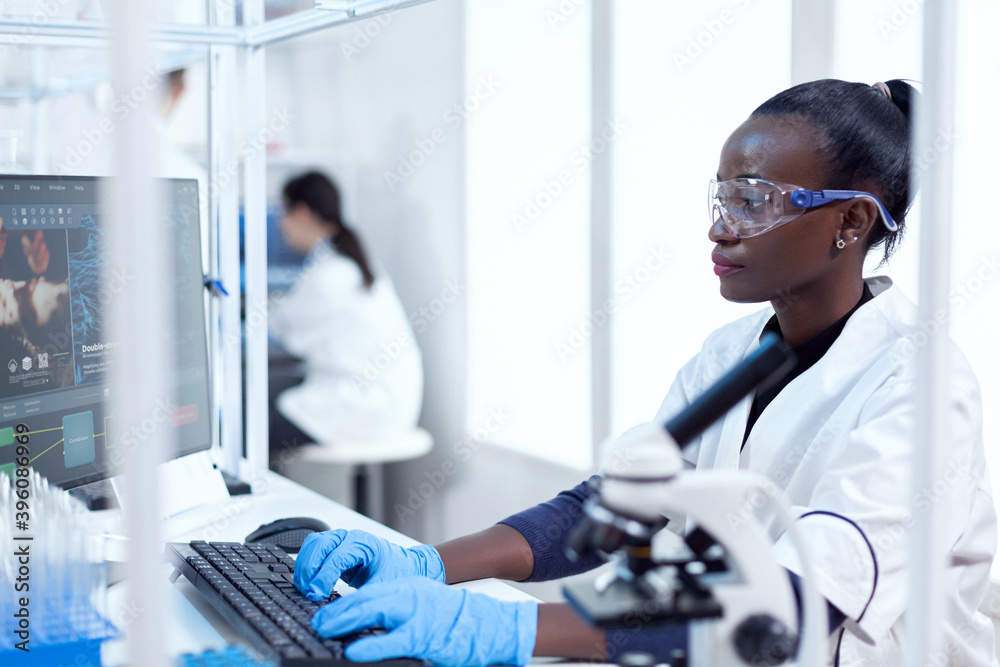 Professional african medical worker typing information about virus study Black healthcare scientist in biochemistry laboratory wearing sterile equipment. Professional african medical worker typing information about virus study Black healthcare scientist in biochemistry laboratory wearing sterile equipment.