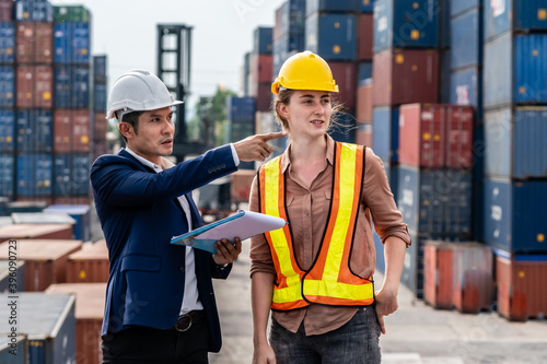 Caucasian woman talking to Asian male manager in a large container yard Wear a safety helmet and reflective vest. for safety Point your finger to the side.