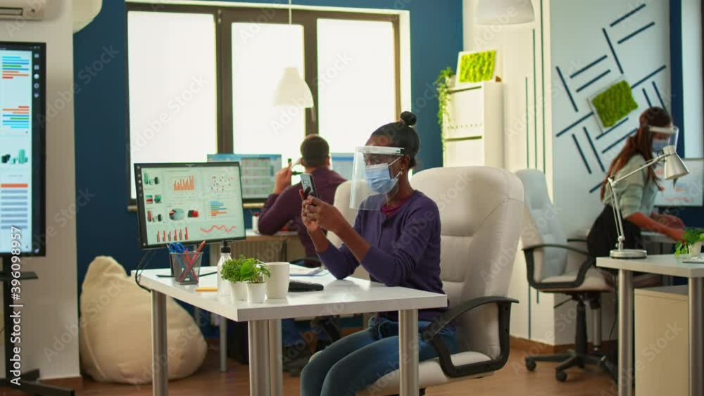 Black businesswoman browsing using smartphone sitting in office room wearing face mask and visor while team doing stategy. Multiethnic coworkers working respecting social distance in financial company Black businesswoman browsing using smartphone sitting in office room wearing face mask and visor while team doing stategy. Multiethnic coworkers working respecting social distance in financial company