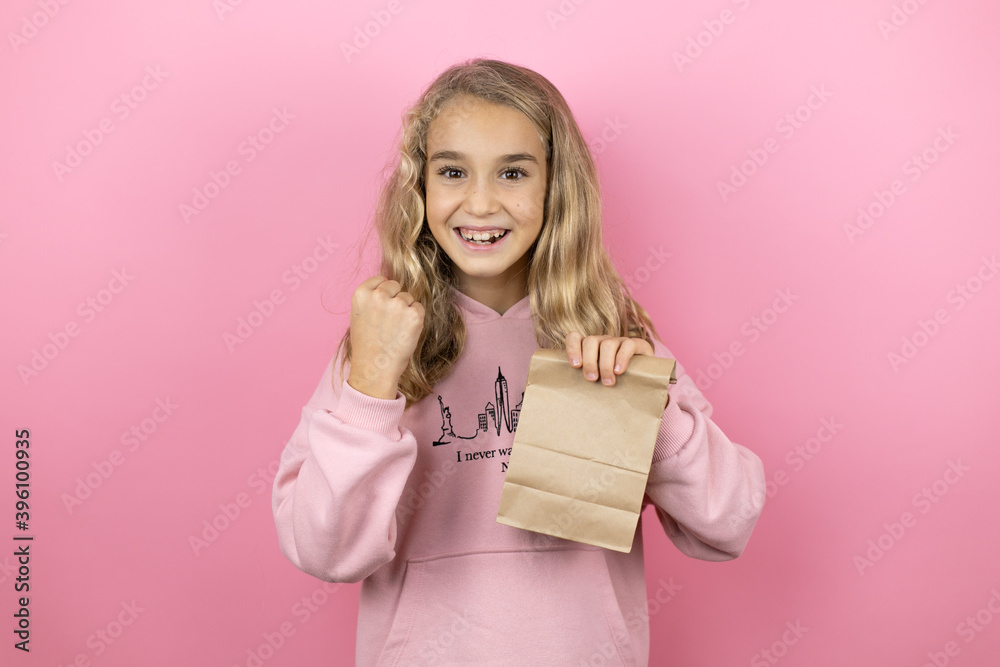 Young beautiful child girl standing over isolated pink background holding a paper bag very happy and excited making winner gesture with raised arms, smiling and screaming for success