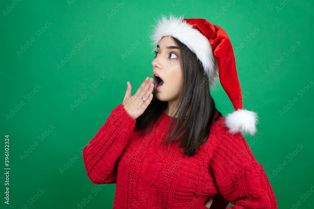 Young beautiful woman wearing Christmas Santa hat over isolated green background with her hand over her mouth and surprised, looking side