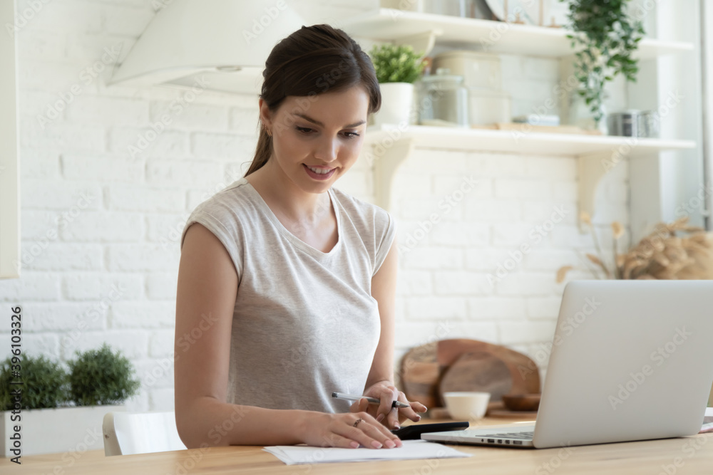 Smiling young woman checking finances, using laptop, sitting at table in kitchen, happy beautiful female calculating domestic bills or expenses, planning budget, working online at home Smiling young woman checking finances, using laptop, sitting at table in kitchen, happy beautiful female calculating domestic bills or expenses, planning budget, working online at home