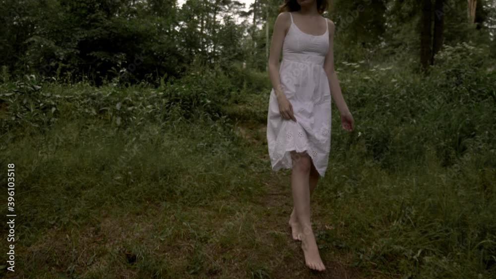 A barefoot lady in a white summer dress with a Daisy flower in her hands descends from a hill in a pine park A barefoot lady in a white summer dress with a Daisy flower in her hands descends from a hill in a pine park