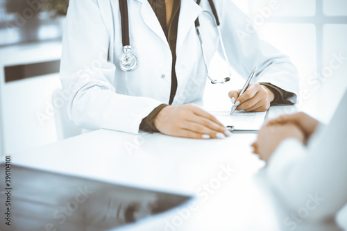 Unknown woman-doctor and female patient sitting and talking at medical examination in clinic, close-up. Therapist wearing green blouse is filling up medication history record. Medicine concept
