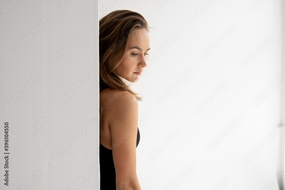 Portrait of young brunette skinny woman is posing in black underwear and standing behind a white wall in white studio. Model tests of pretty girl in basic clothes on cyclorama. Portrait of young brunette skinny woman is posing in black underwear and standing behind a white wall in white studio. Model tests of pretty girl in basic clothes on cyclorama.