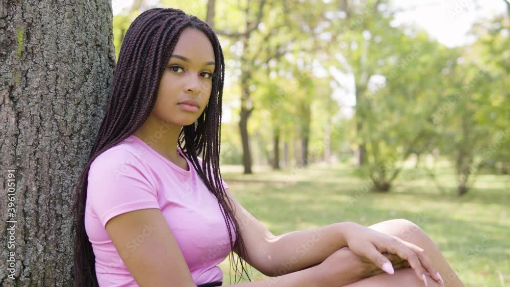 A young black woman looks seriously at the camera as she sits under a tree in a park on a sunny day