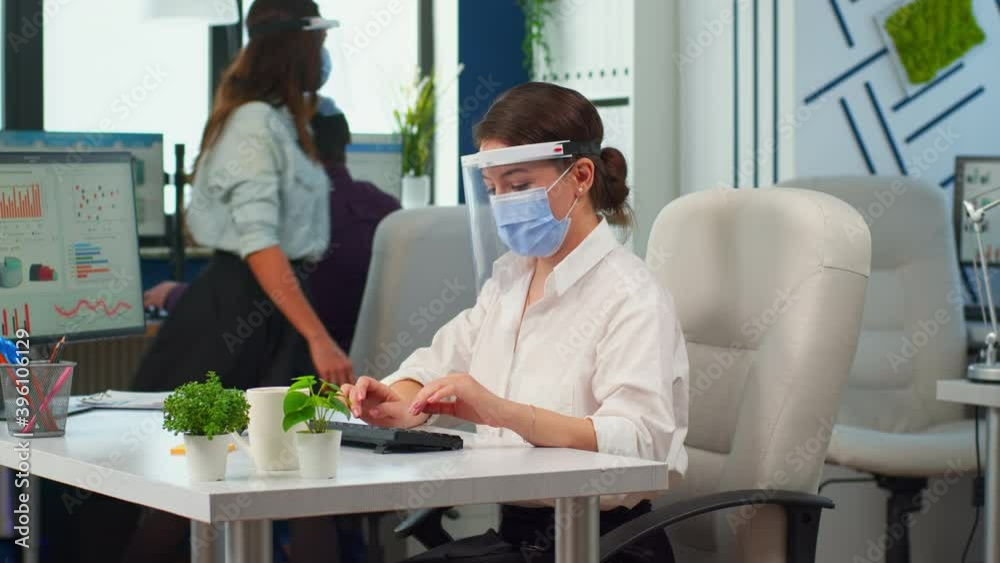 Company manager with face mask typing on computer and looking on camera in new normal business office respecting social distance during coronavirus pandemic. Colleagues working in background. Company manager with face mask typing on computer and looking on camera in new normal business office respecting social distance during coronavirus pandemic. Colleagues working in background.