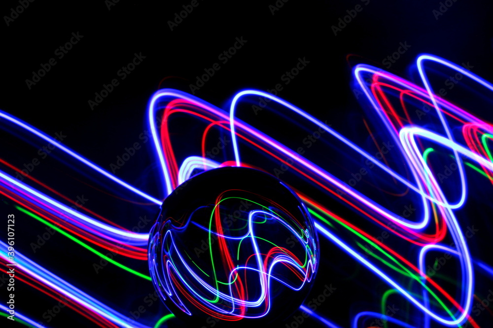 Long exposure photograph of neon multi colour in an abstract swirl, parallel lines pattern against a black background with reflections in a glass crystal ball. Light painting photography.