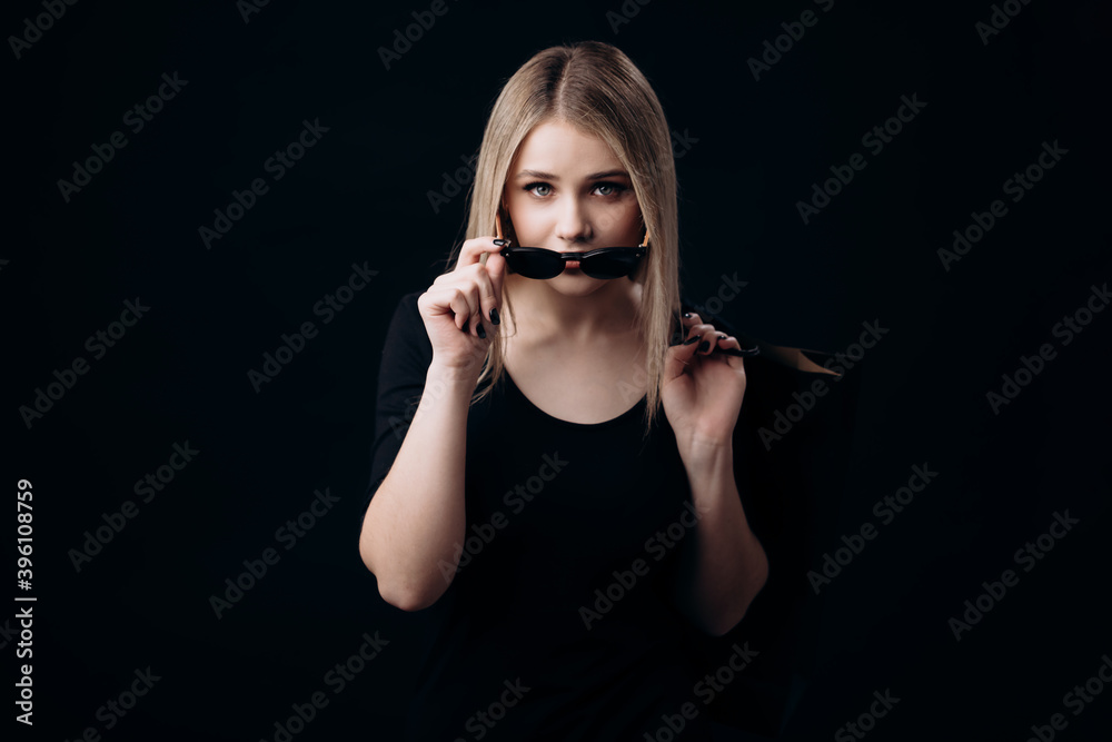 Fototapeta premium Front view of charming young woman wearing sunglasses, standing over black background and holding shopping bags. Black friday concept.
