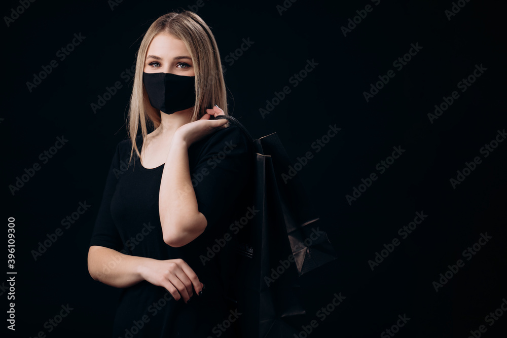 Obraz premium Pretty young lady in medical protective mask holding paper bags over black studio background. Concept of sales and purchase.