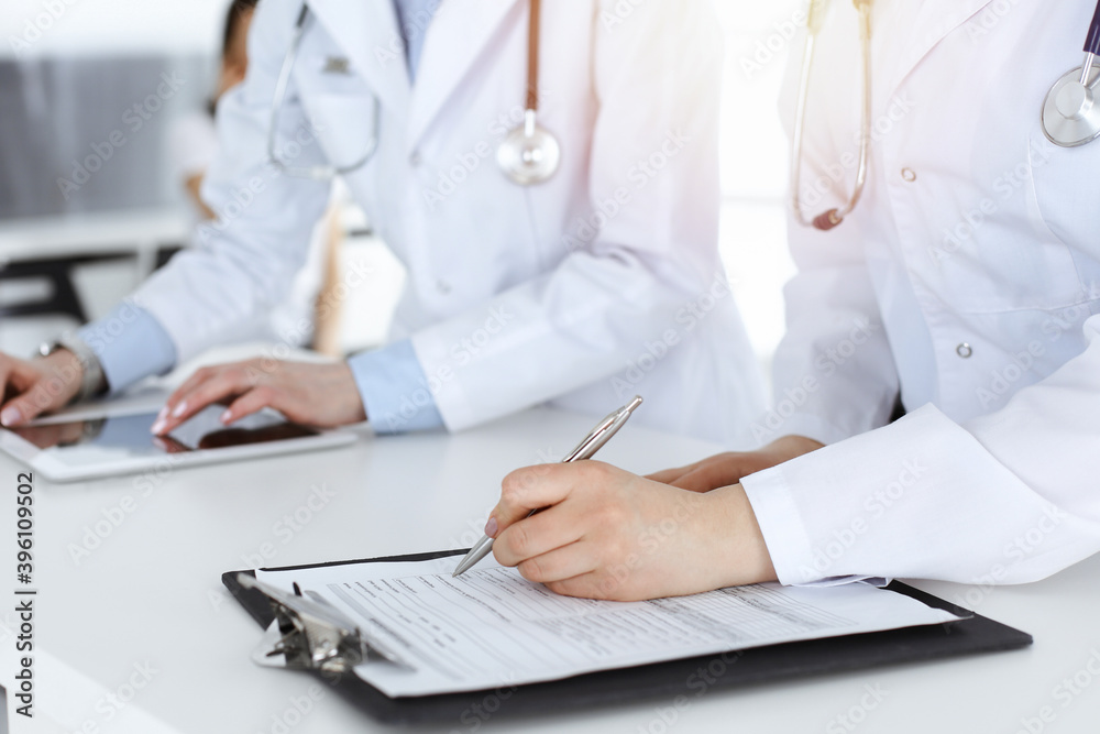 Unknown woman-doctors at work with patient at the background. Female physicians filling up medical documents or prescription while standing in hospital reception desk, close-up. Health care concept Unknown woman-doctors at work with patient at the background. Female physicians filling up medical documents or prescription while standing in hospital reception desk, close-up. Health care concept