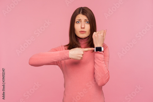 Displeased impatient woman with brown hair in pink sweater pointing finger on her wrist clock looking at camera with anxious, worried about deadline. Indoor studio shot isolated on pink background