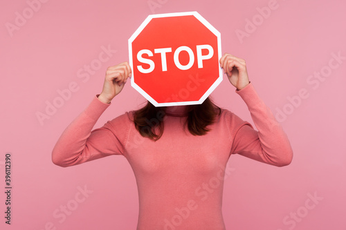 Brunette woman in pink sweater hiding face behind stop sign, way prohibited, stop violence. Indoor studio shot isolated on pink background