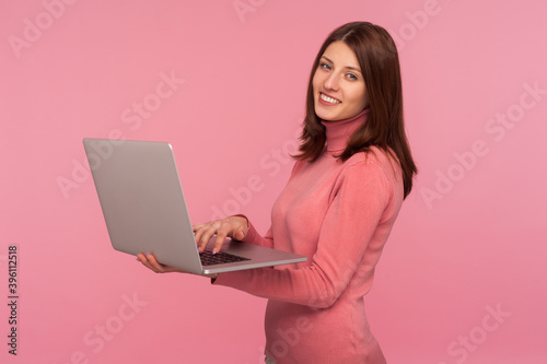 Happy positive woman with brown hair in pink sweater working on laptop and looking at camera with toothy smile, enjoying remote work. Indoor studio shot isolated on pink background