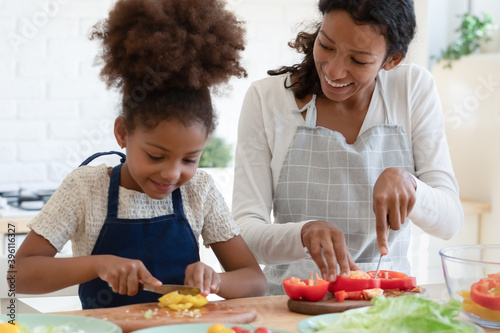 Culinary is easy. Inspired happy african american mother and tween daughter prepare vegan food  grown elder and small younger sisters trying new cooking recipe  cutting vegetables talking at kitchen