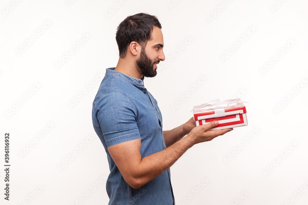 Profile portrait smiling bearded man in blue shirt making present holding in hands striped gift box, celebrating anniversary, preparing for holidays. Indoor studio shot isolated on white background Profile portrait smiling bearded man in blue shirt making present holding in hands striped gift box, celebrating anniversary, preparing for holidays. Indoor studio shot isolated on white background