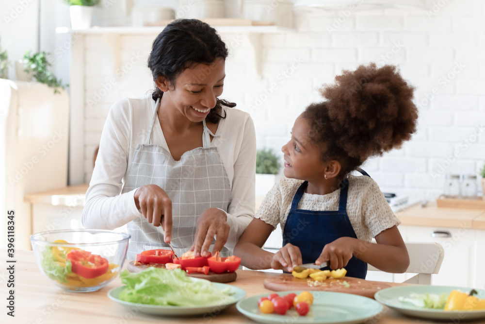 Cooking lesson. Happy patient black foster mom teaching little adopted daughter prepare healthy nutrition, single african mommy and preteen girl talking smiling engaged in useful activity at kitchen