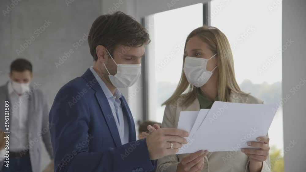Young man and woman with protective facial masks discussing with paper in hands indoors in the office with young people works behind them Young man and woman with protective facial masks discussing with paper in hands indoors in the office with young people works behind them