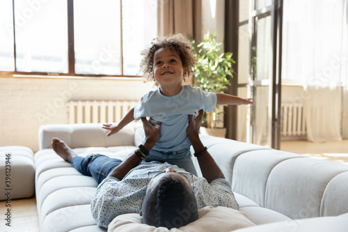 Having fun together. Excited laughing black foster father and little adopted son resting playing on cozy sofa, cute small african kid imagining himself an airplane being raised up to air in dad hands