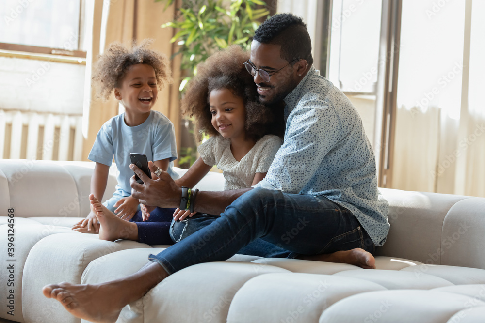 Funny movie. Caring millennial african american foster father and two little adopted kids black brother and sister having fun at home sitting on sofa close together watching cute cartoons on cellphone Funny movie. Caring millennial african american foster father and two little adopted kids black brother and sister having fun at home sitting on sofa close together watching cute cartoons on cellphone