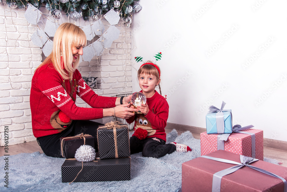 Young attractive smiling woman with her daughter considering a Christmas gift near the fireplace with Christmas decor. Place to write or greet.